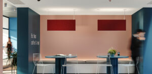 Two red Rail 2 Acoustic luminaires hang in an off lounge area both lights showing illumination above on the ceiling and below on the tables